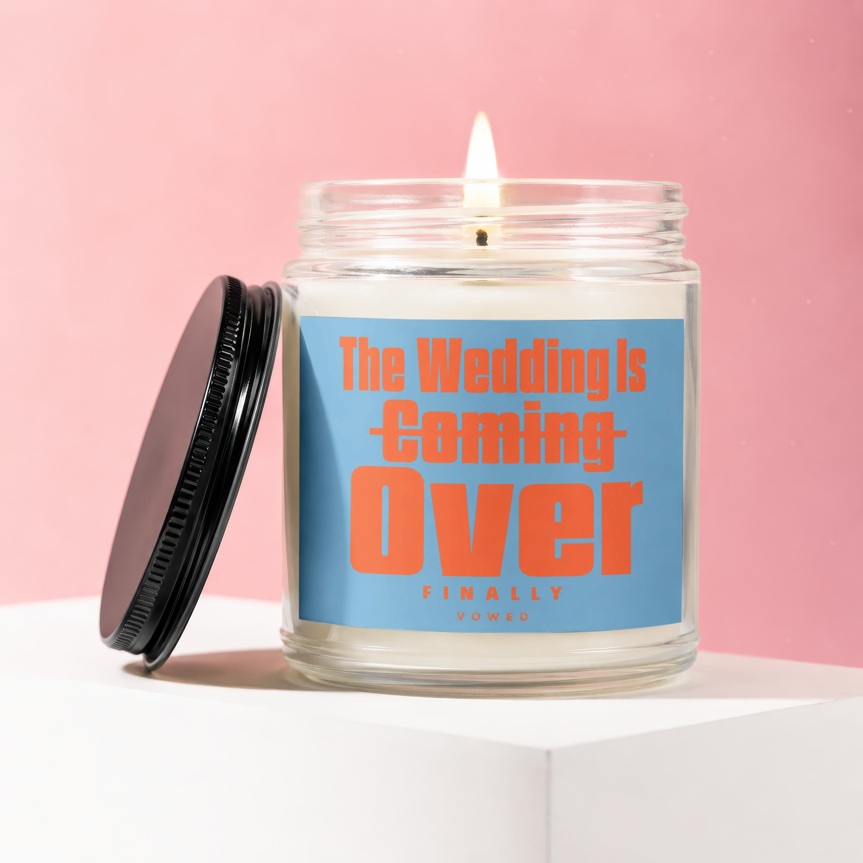 Lit candle in glass jar with black lid, reads "The Wedding Is Coming Over FINALLY VOWED".