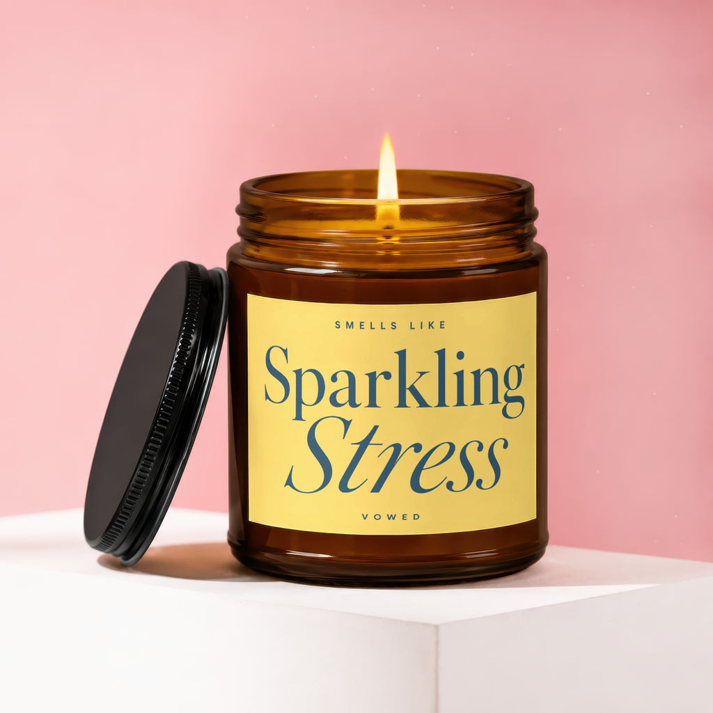 Burning amber jar candle with yellow label reading "Smells Like Sparkling Stress."