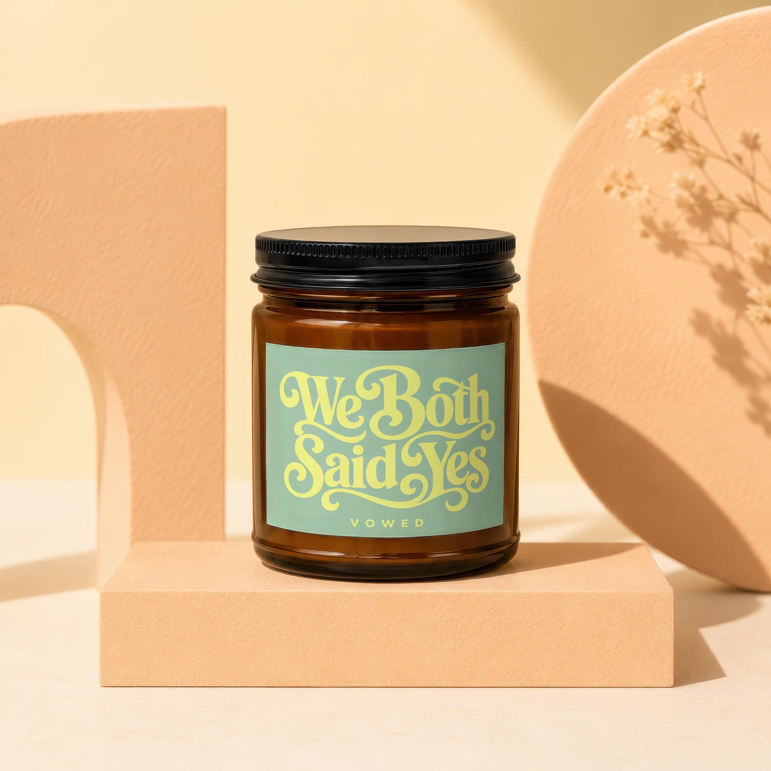 Amber jar candle with "We Both Said Yes VOWED" text in light green script.