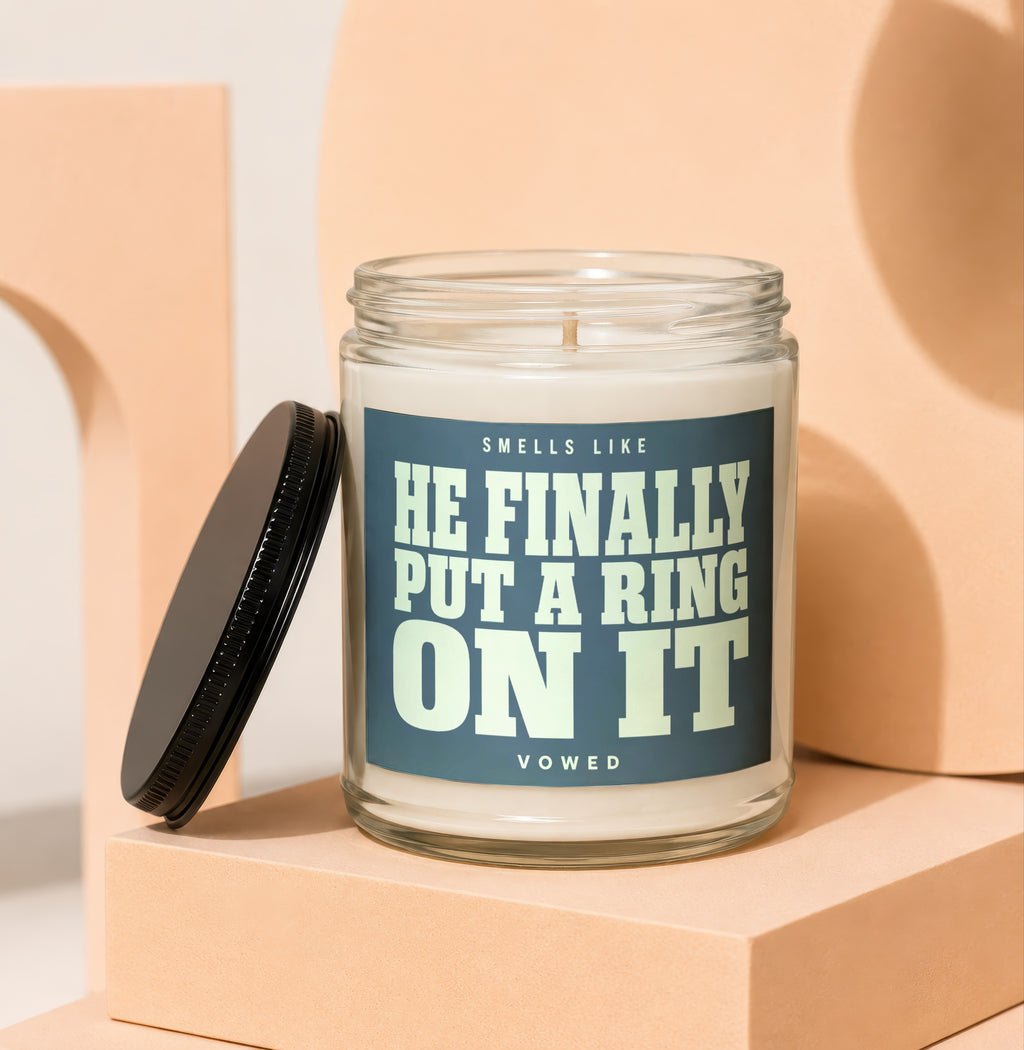 Glass candle with dark blue label reading "SMELLS LIKE HE FINALLY PUT A RING ON IT VOWED".