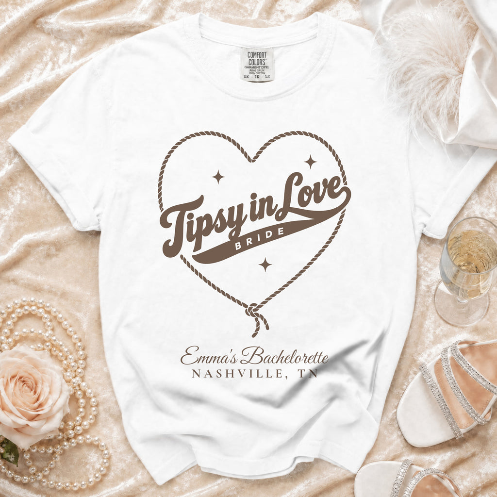 White t-shirt with brown rope heart graphic, Tipsy in Love text, and Emma's Bachelorette Nashville, TN text.