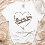 White t-shirt with brown rope heart graphic, Tipsy in Love text, and Emma's Bachelorette Nashville, TN text.