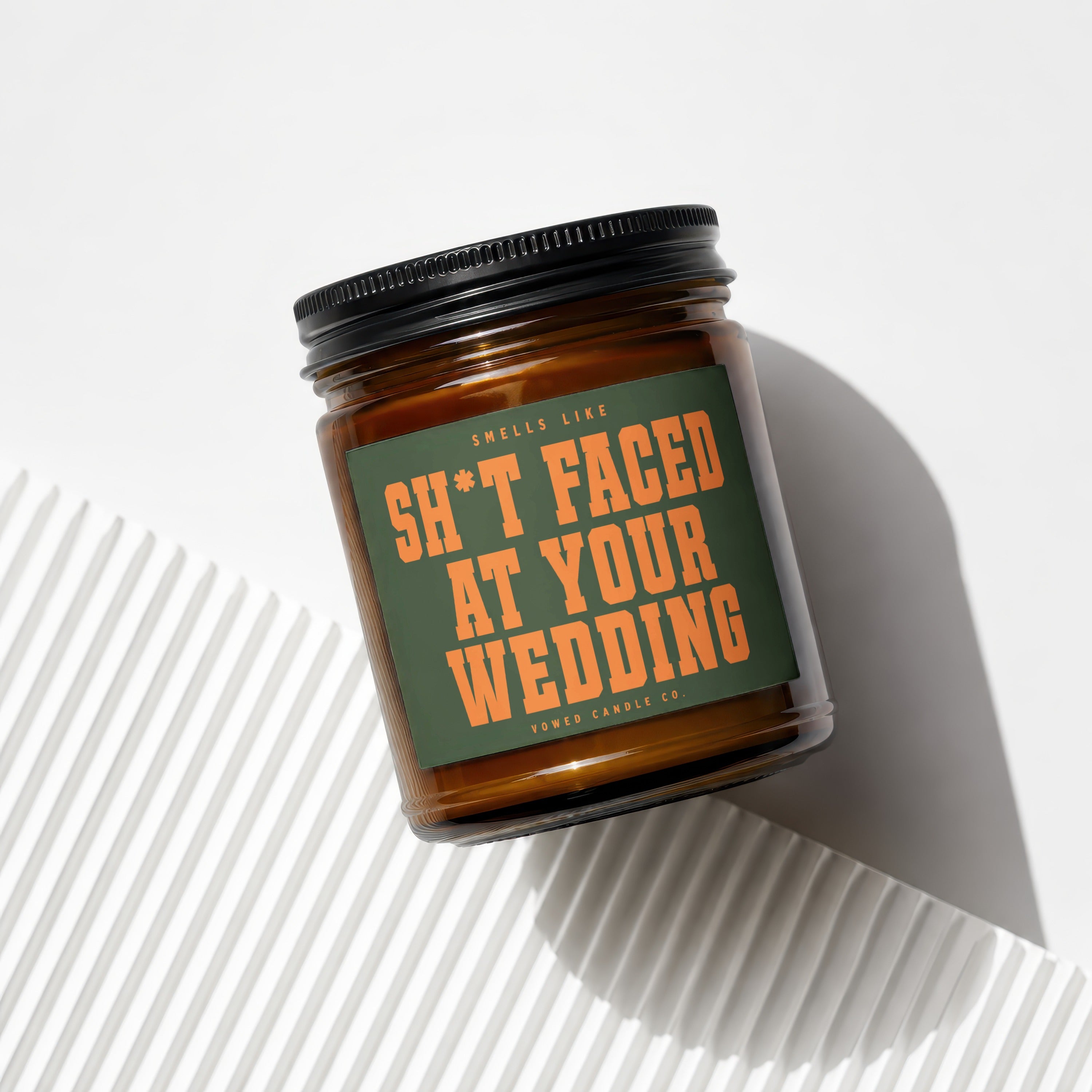 Amber candle in glass jar with black lid, green label reads "Smells like Sh*t Faced At Your Wedding"