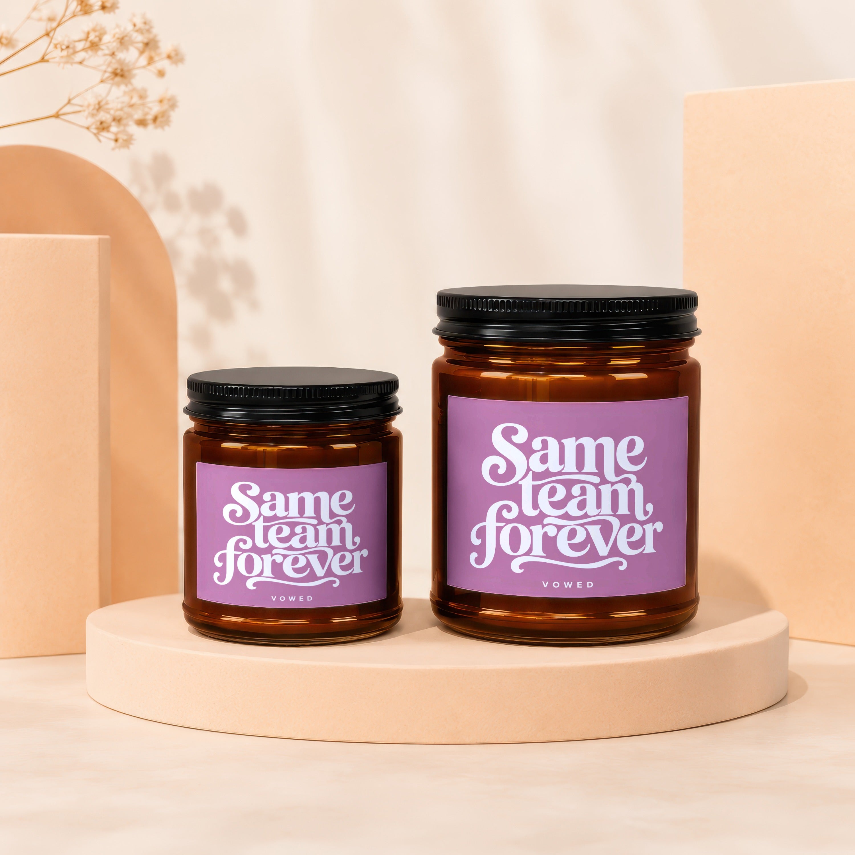 Two amber glass candles, one larger than the other, with purple labels reading "Same team forever".