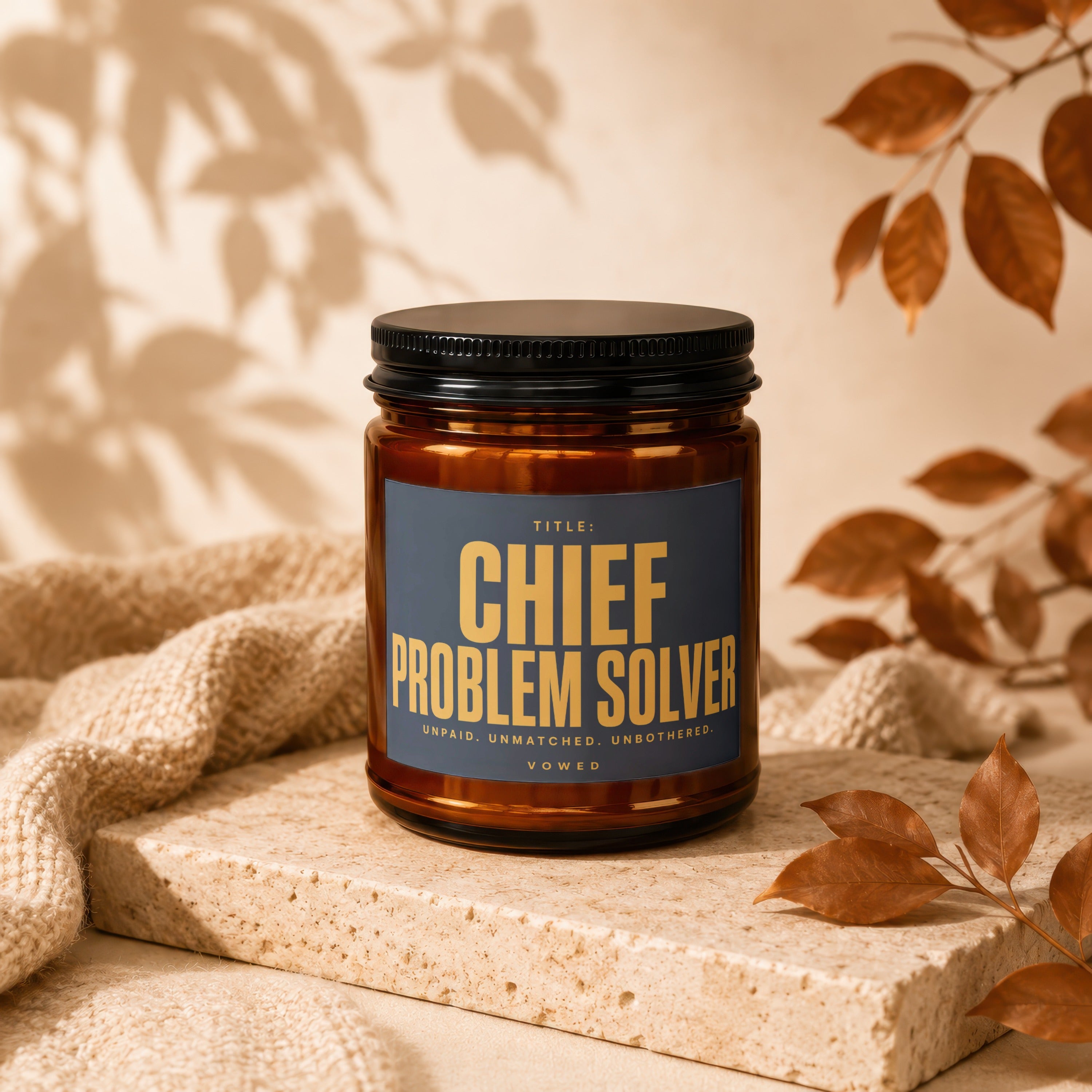 Brown candle with "Chief Problem Solver" label and descriptive text, with autumn leaves and fabric backdrop.