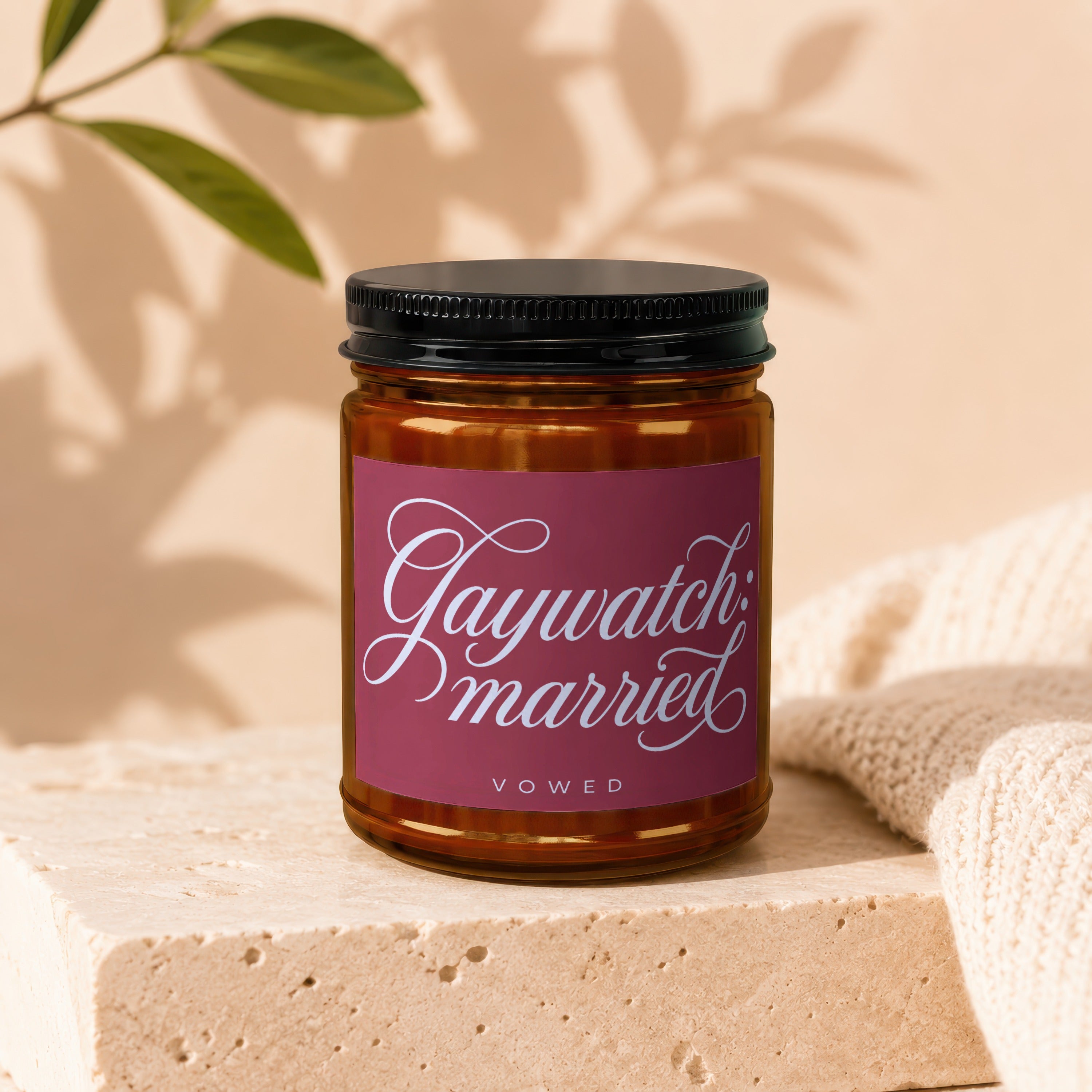 Candle with "Gaywatch married" and "vowed" text on label, framed by light and a textured knit.