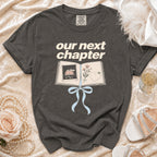 Dark gray t-shirt with "our next chapter" text and open book illustration.