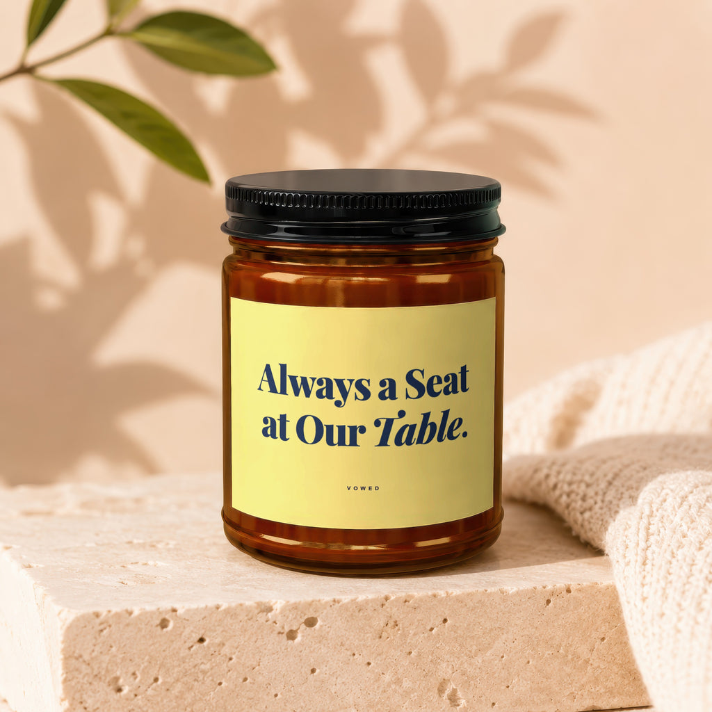 Candle with black lid, yellow label text "Always a Seat at Our Table. VOWED."