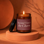 Burning amber candle with lit wick on orange pedestal, showing text "Second Time's the Charm".