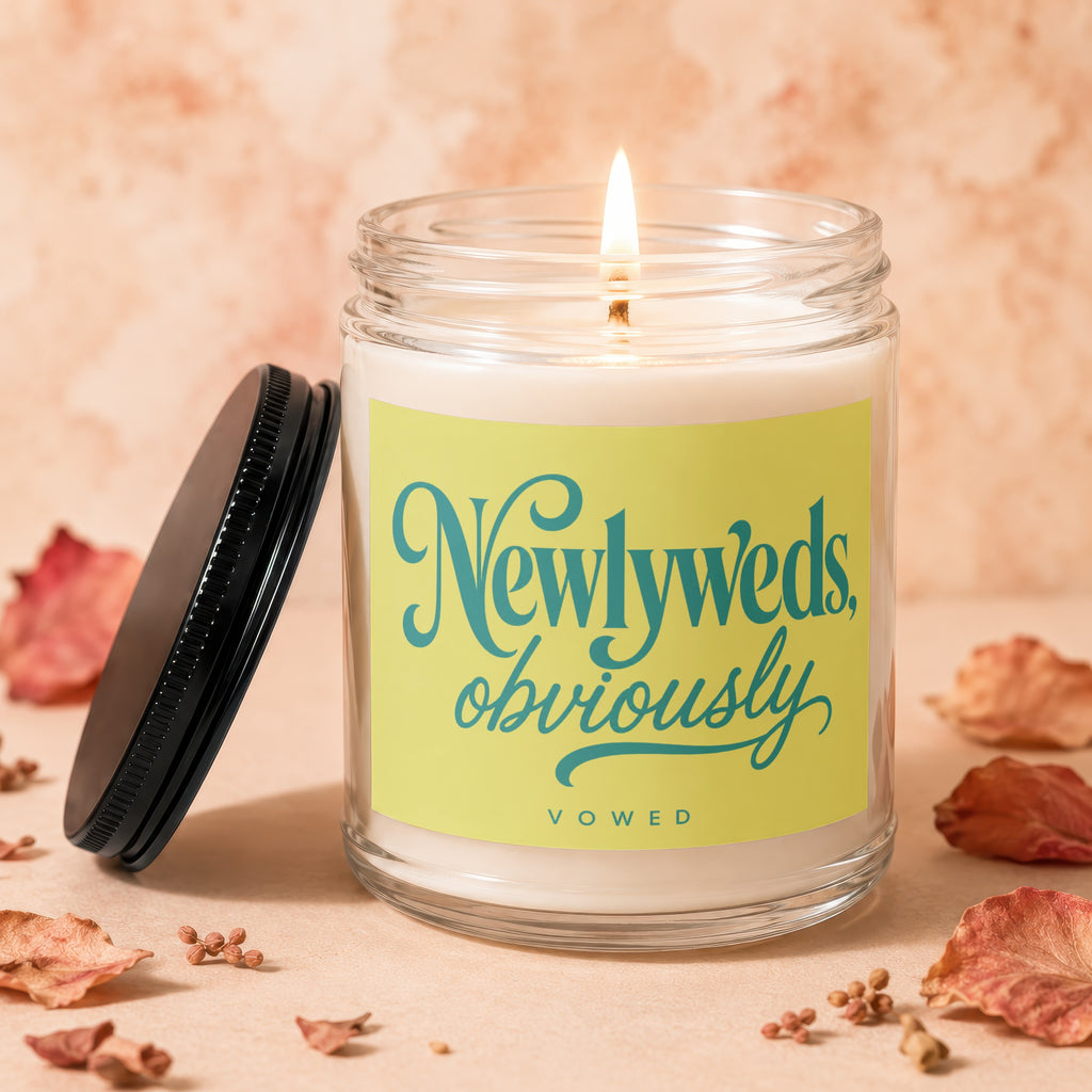 Glass jar candle with a lit wick and a yellow label reading "Newlys, obviously, vowed".