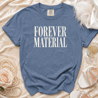 Blue t-shirt with "FOREVER MATERIAL" text, laid flat with decorative accessories.