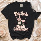 Black t-shirt with "This Bride Needs Champagne" text, bottle, and Champagne glass graphic.