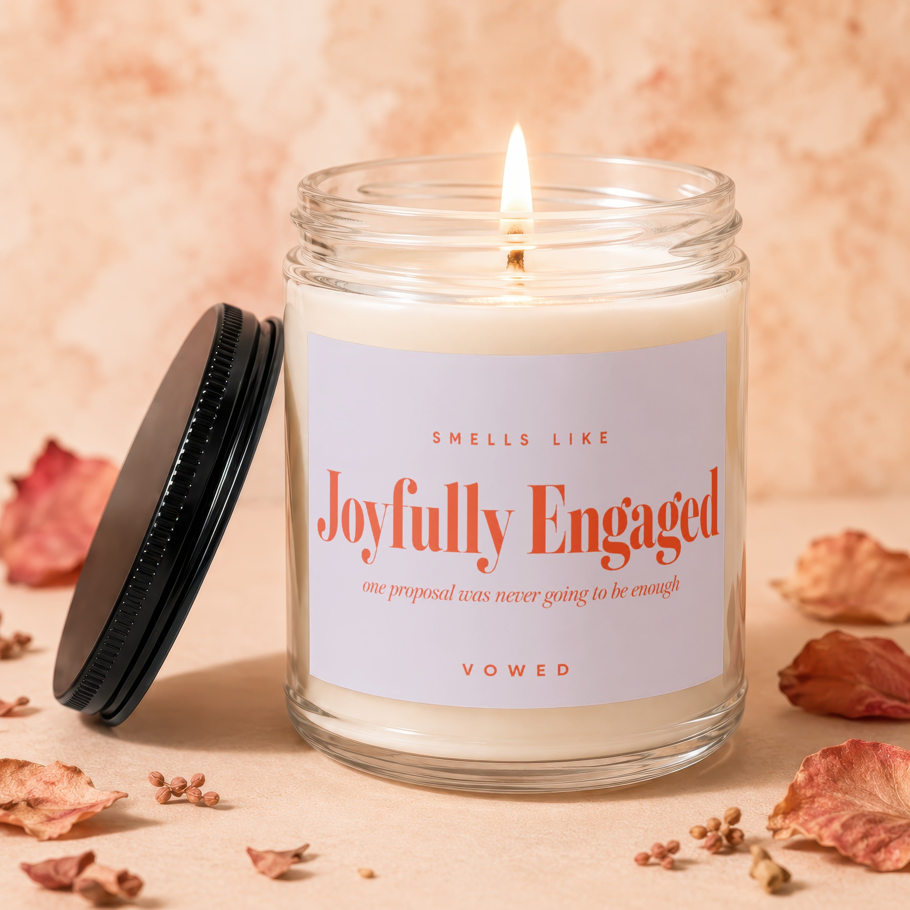 A lit candle in a glass jar labeled "Joyfully Engaged" with decorative dried leaves scattered.