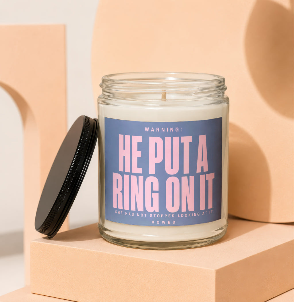 Glass candle with blue label that reads "WARNING: HE PUT A RING ON IT" and smaller text.