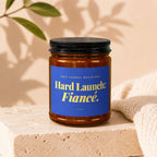 Amber jar candle with blue label reading "Hard Launch: Fiancé." and "Vowed."