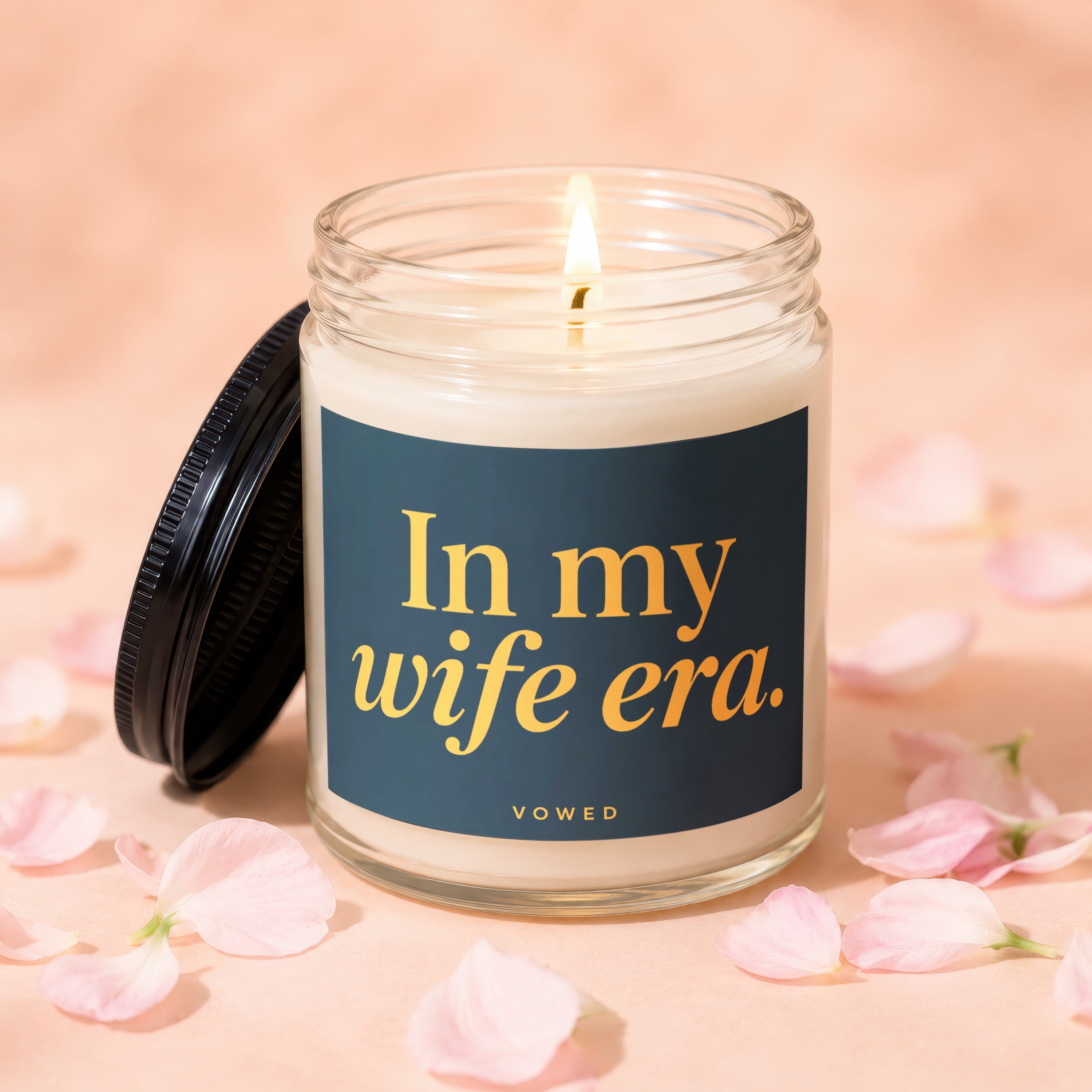 Lit glass candle with dark label reading "In my wife era." surrounded by pink petals.