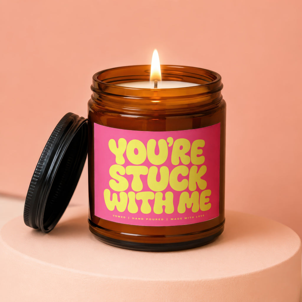 Amber jar candle with lit flame and pink label reading "YOU'RE STUCK WITH ME".