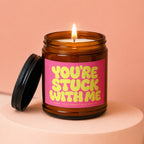 Amber jar candle with lit flame and pink label reading "YOU'RE STUCK WITH ME".