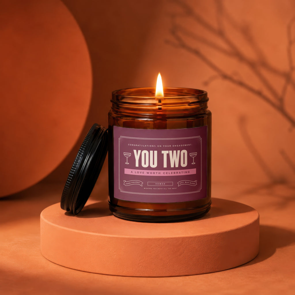 Amber jar candle with lit wick, label reads "YOU TWO A LOVE WORTH CELEBRATING."