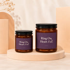 Two amber glass candles with purple labels that read "Ring On, Heart Full."