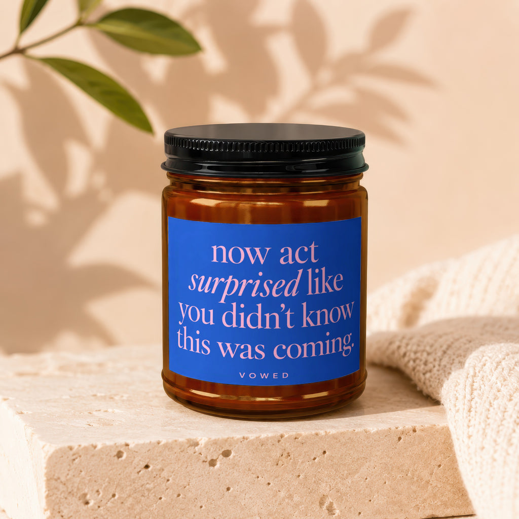 Amber jar candle with blue label reading "now act surprised like you didn't know this was coming."