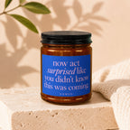 Amber jar candle with blue label reading "now act surprised like you didn't know this was coming."