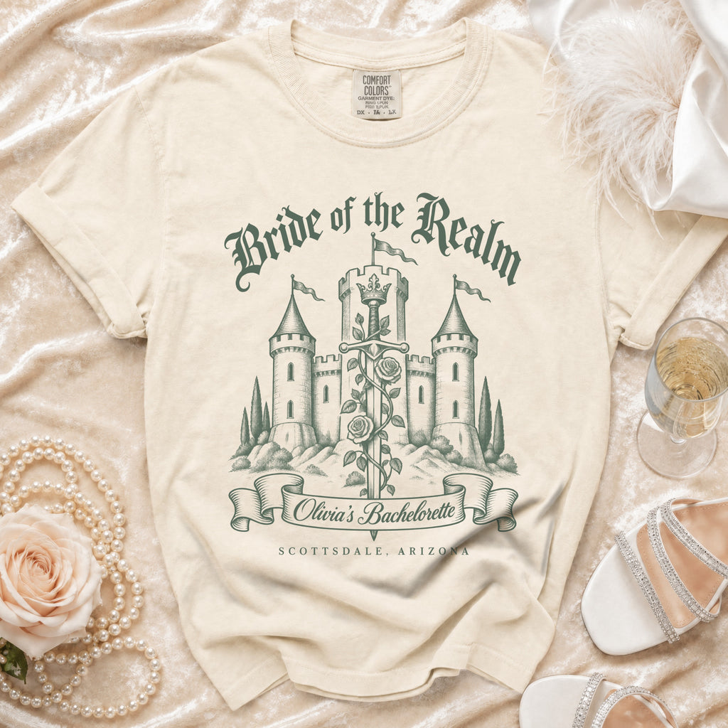 A cream-colored t-shirt with a green castle graphic, sword, roses, and text reading "Bride of the Realm Olivia's Bachelorette Scottsdale, Arizona".