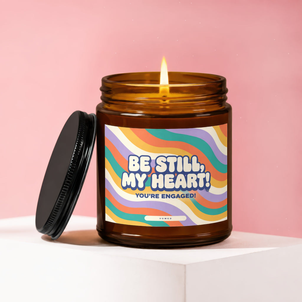 Amber candle with lit wick, black lid, and label that reads "BE STILL, MY HEART! YOU'RE ENGAGED!"