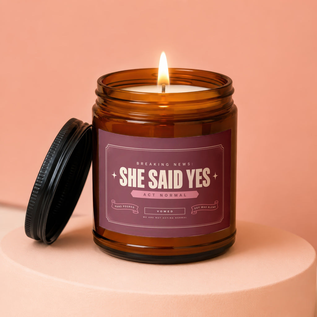 Amber glass candle labeled "She Said Yes" with a lit wick and black lid.
