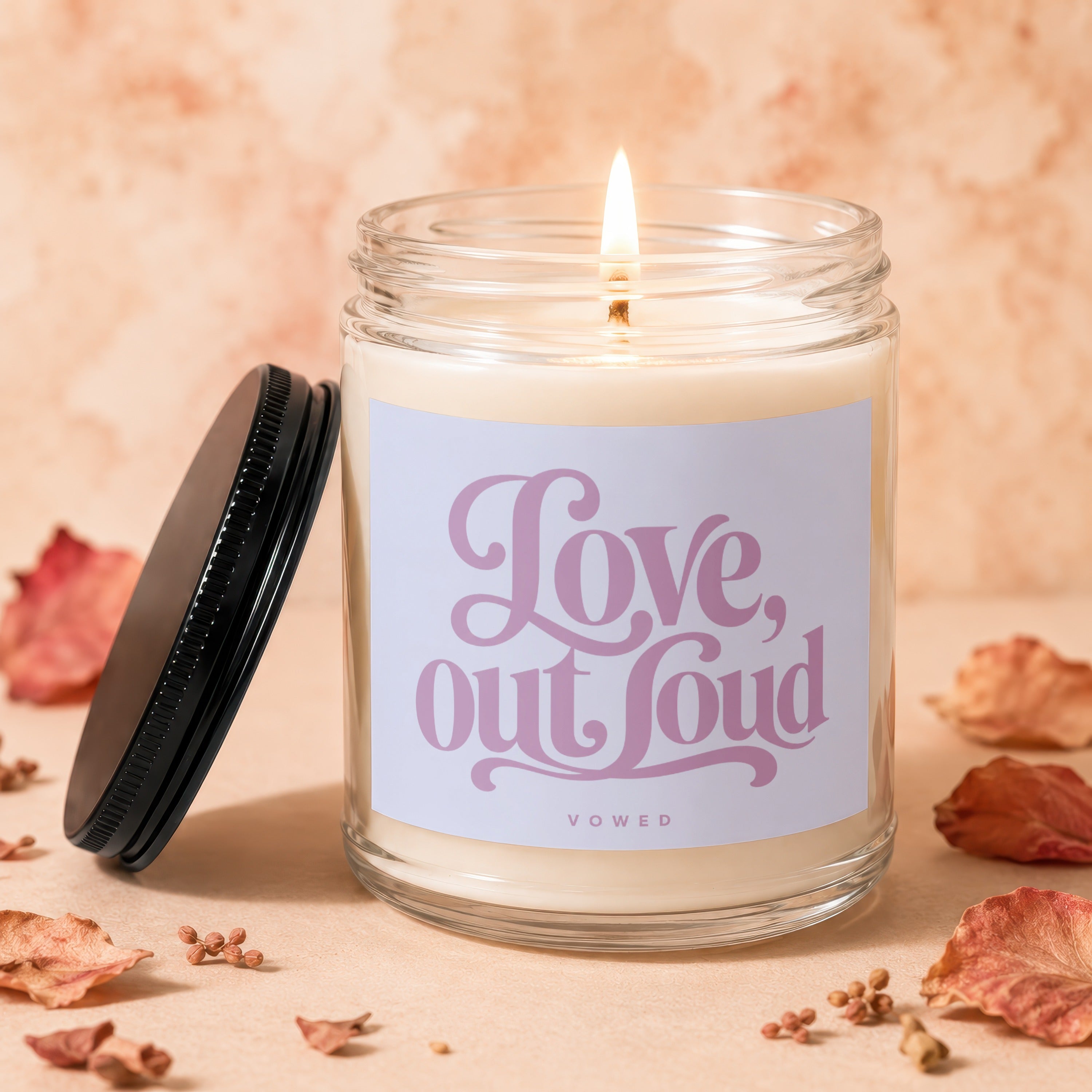 Lit candle in glass jar with "Love, out loud" text and "VOWED" brand.