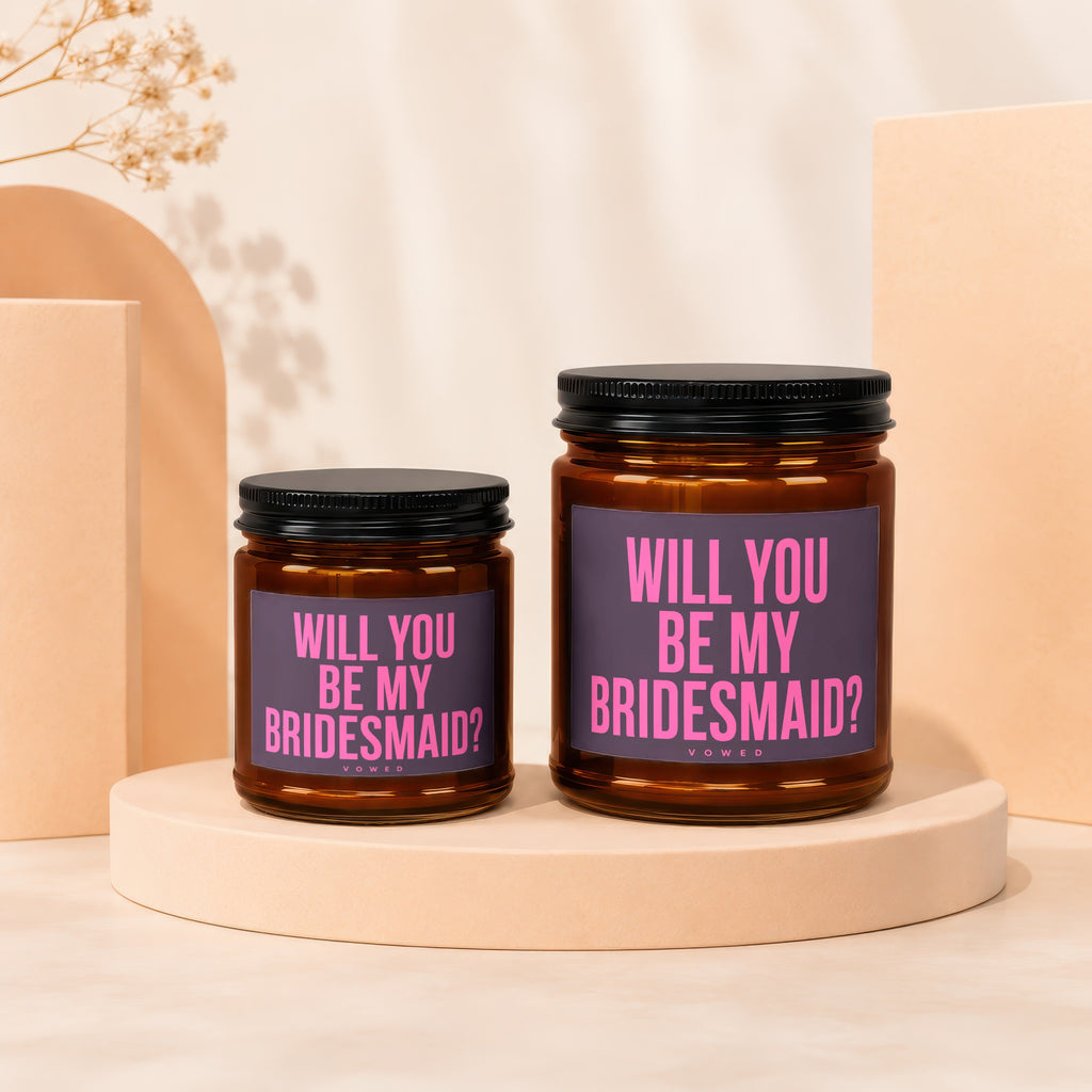 Two amber glass candles with black lids sit on beige pedestals, reading "Will You Be My Bridesmaid?".