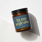 Brown glass candle with black lid, featuring a dark blue label with gold text: "In my wife era."