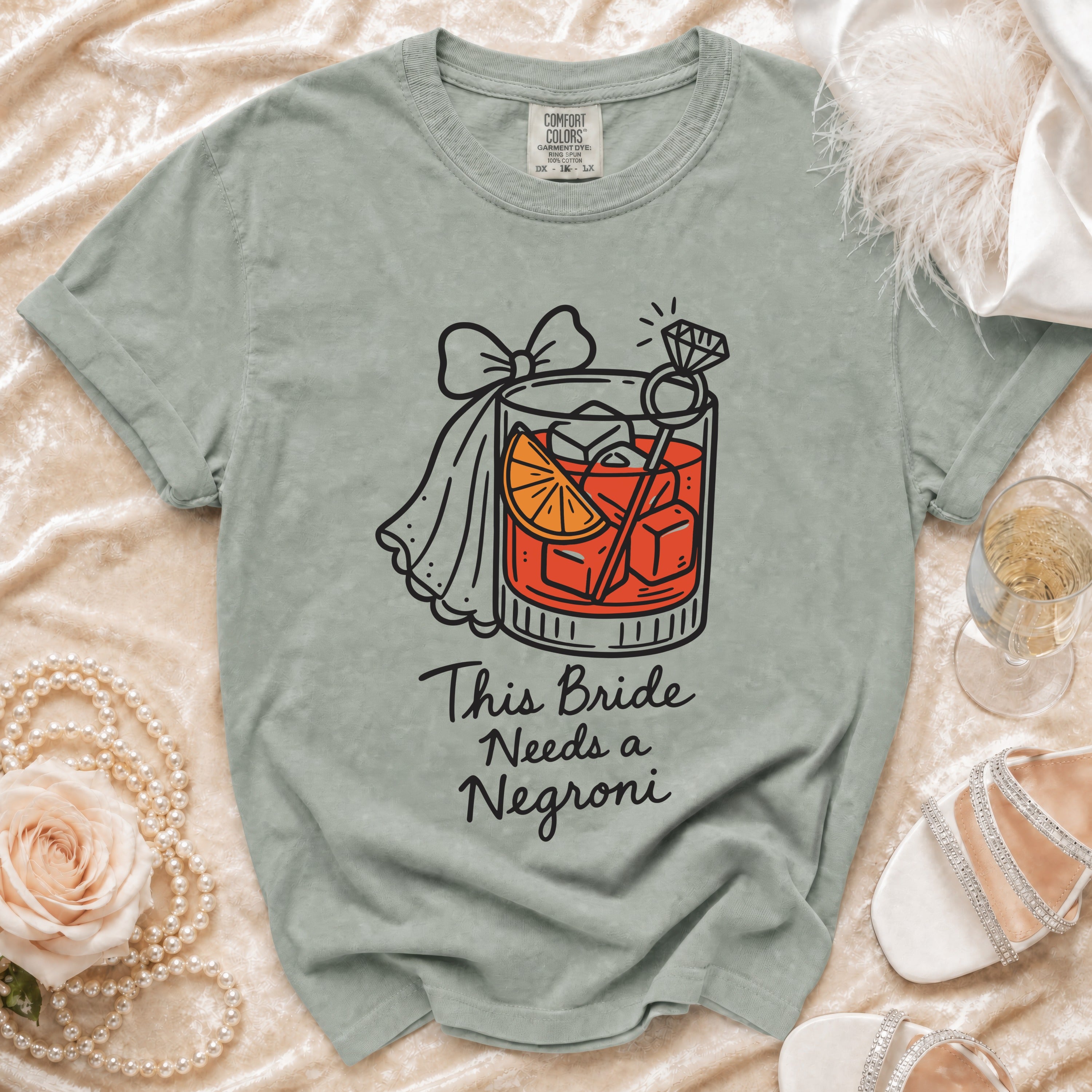 Light green t-shirt with graphic of a negroni, ring, and text "This Bride Needs a Negroni".
