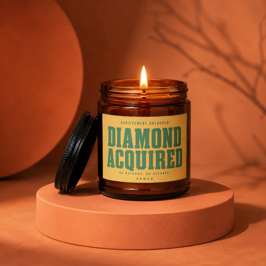 Amber jar candle with "DIAMOND ACQUIRED, VOWED" text, lid open, lit.
