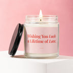 Glass candle with lit wick, black lid, and pink label that reads "Wishing You Luck a Lifetime of Love. VOWED."