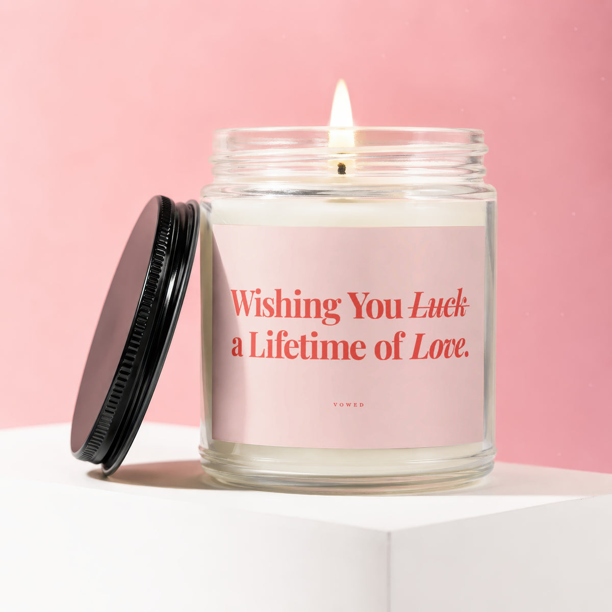 Glass candle with lit wick, black lid, and pink label that reads "Wishing You Luck a Lifetime of Love. VOWED."