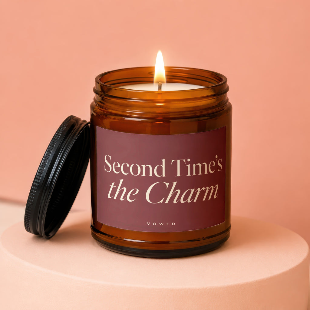 Amber glass candle with burning wick and lid, reads "Second Time's the Charm".
