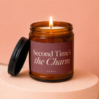 Amber glass candle with burning wick and lid, reads "Second Time's the Charm".