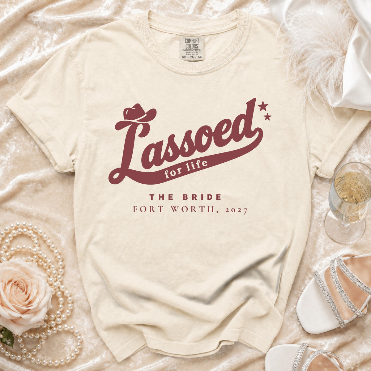 A cream-colored t-shirt displays maroon text reading "Lassoed for life The Bride Fort Worth, 2027."