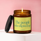 An amber candle with a lit wick and black lid, labeled "The people who showed up"