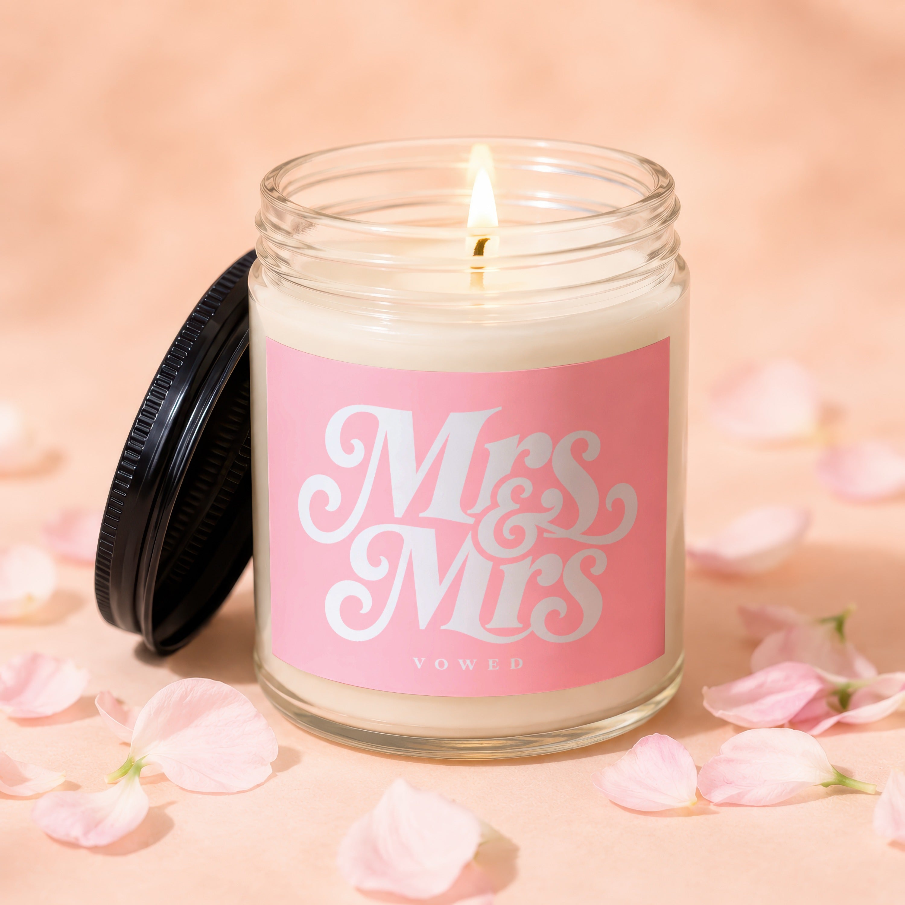 Candle with "Mrs & Mrs Vowed" text on pink label, lid off, surrounded by petals.