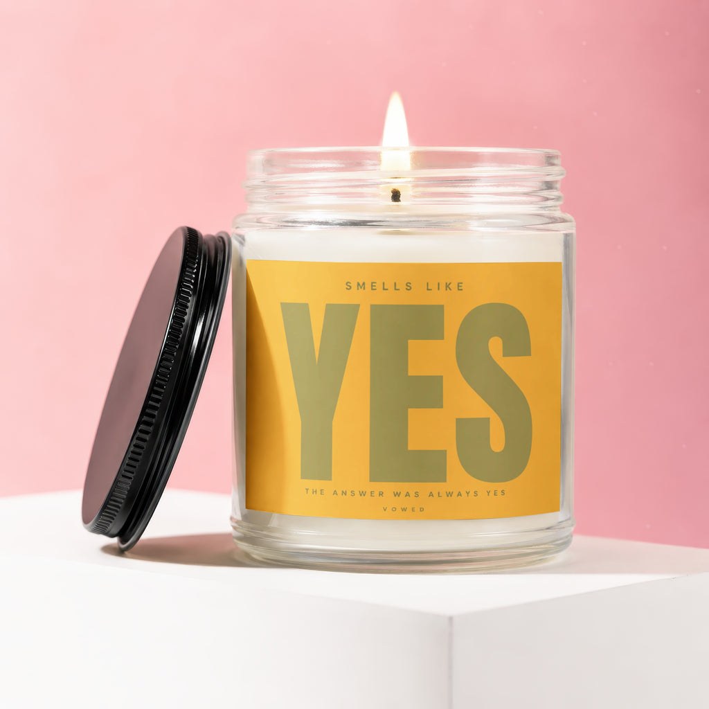 Lit candle in glass jar with yellow label reading "SMELLS LIKE YES THE ANSWER WAS ALWAYS YES"