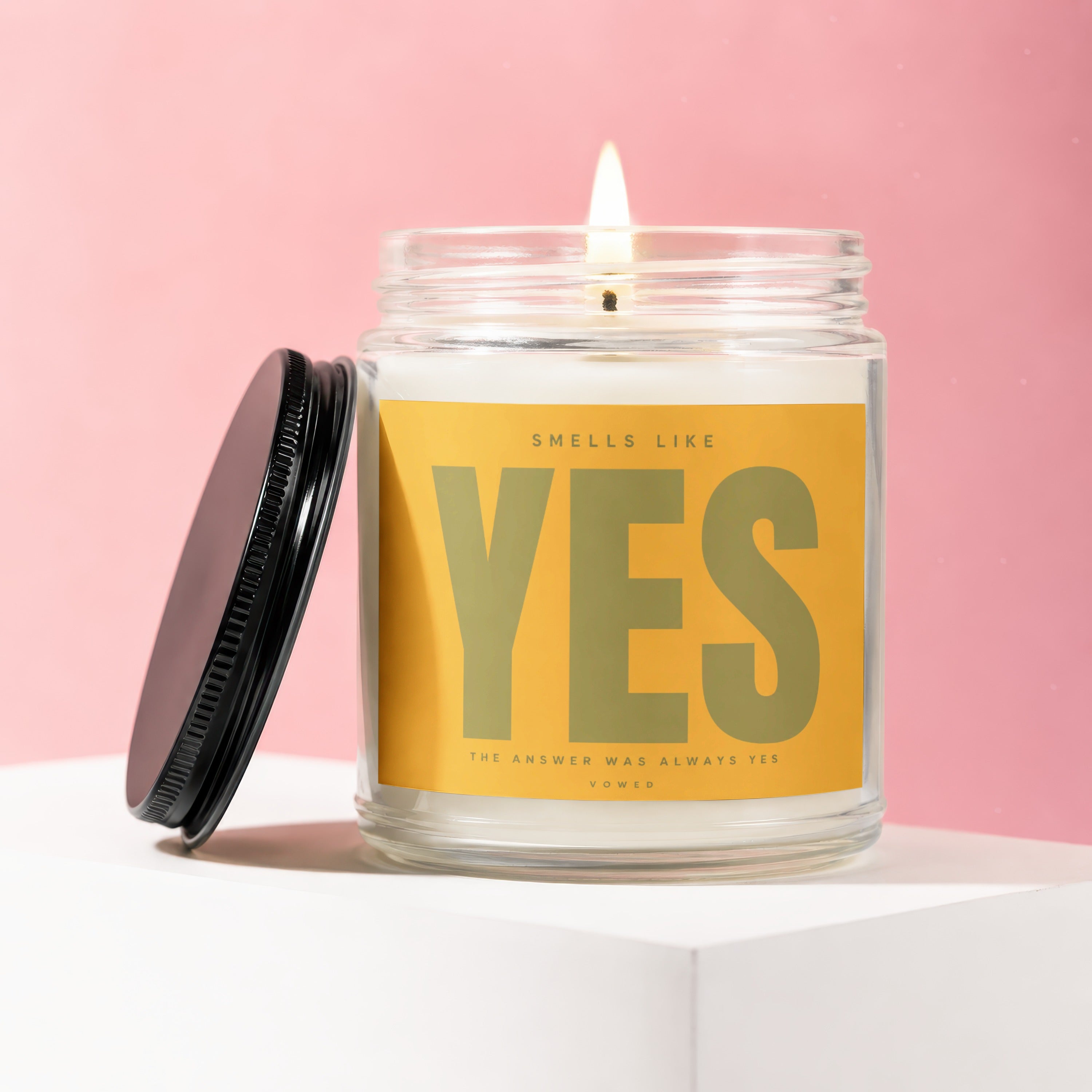 Lit candle in glass jar with yellow label reading "SMELLS LIKE YES THE ANSWER WAS ALWAYS YES"