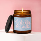 Amber glass candle with a lit wick and a black lid, featuring text "Two brides, one aisle."