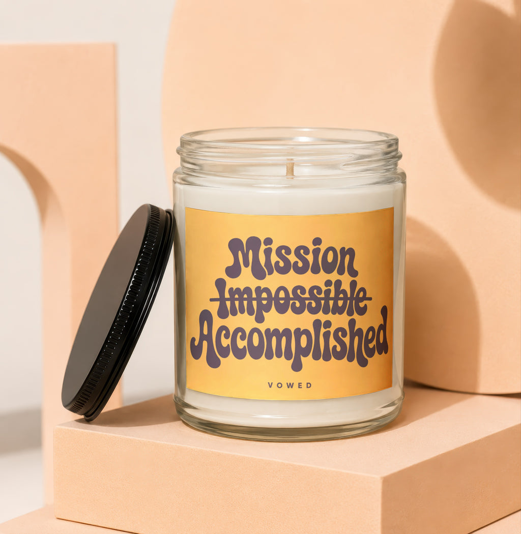 Glass jar candle with yellow label reading "Mission Impossible Accomplished VOWED".