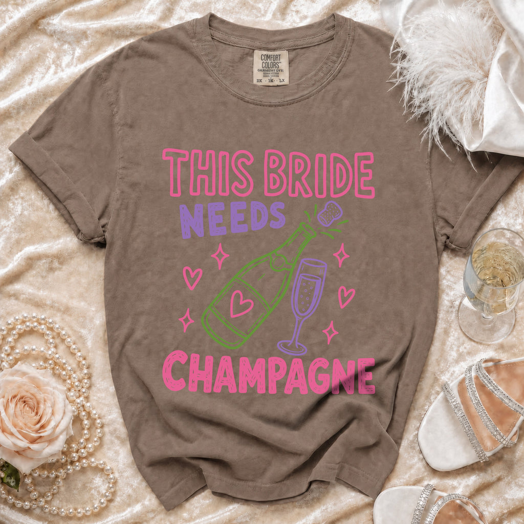 Brown t-shirt with pink and purple text: "THIS BRIDE NEEDS CHAMPAGNE".