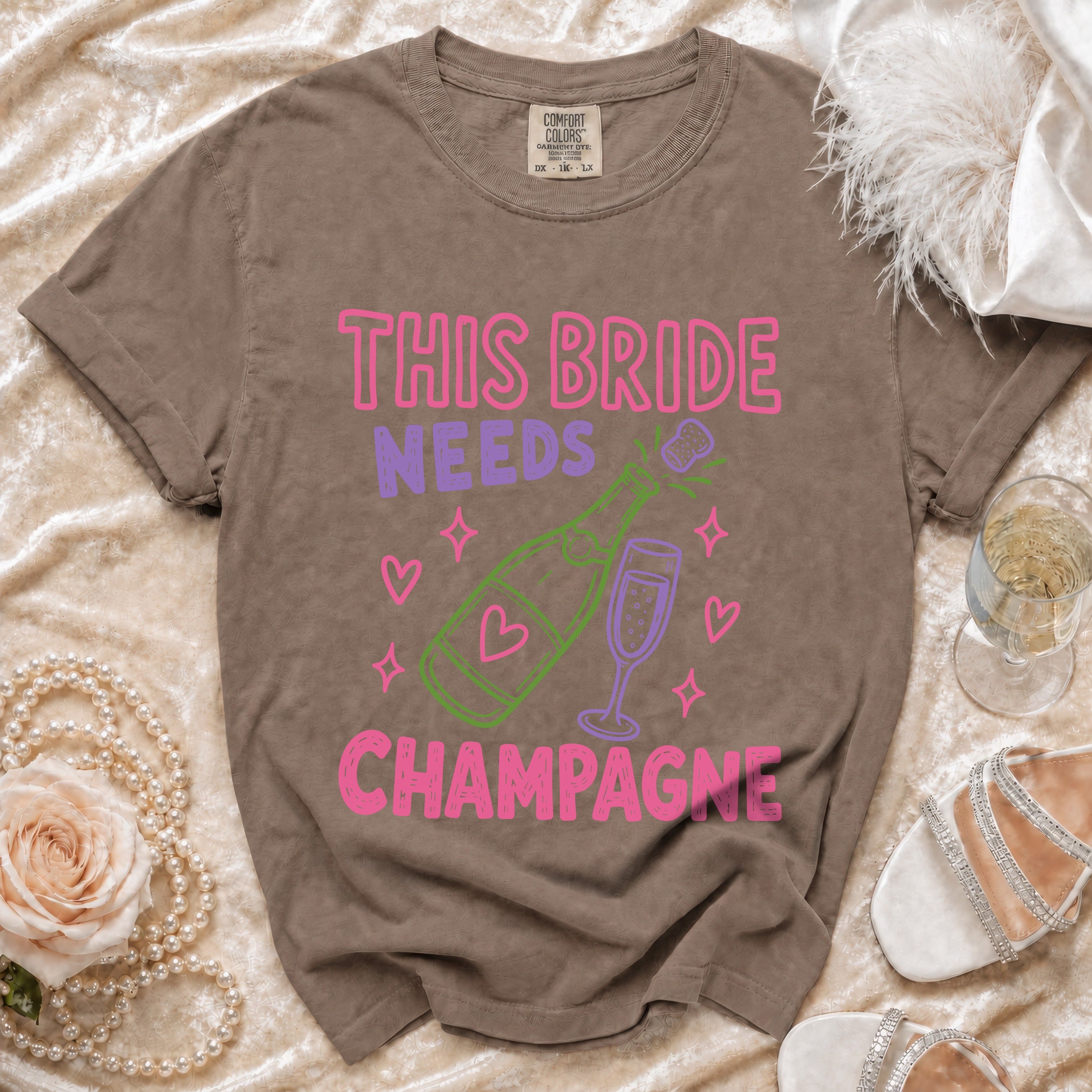 Brown t-shirt with pink and purple text: "THIS BRIDE NEEDS CHAMPAGNE".