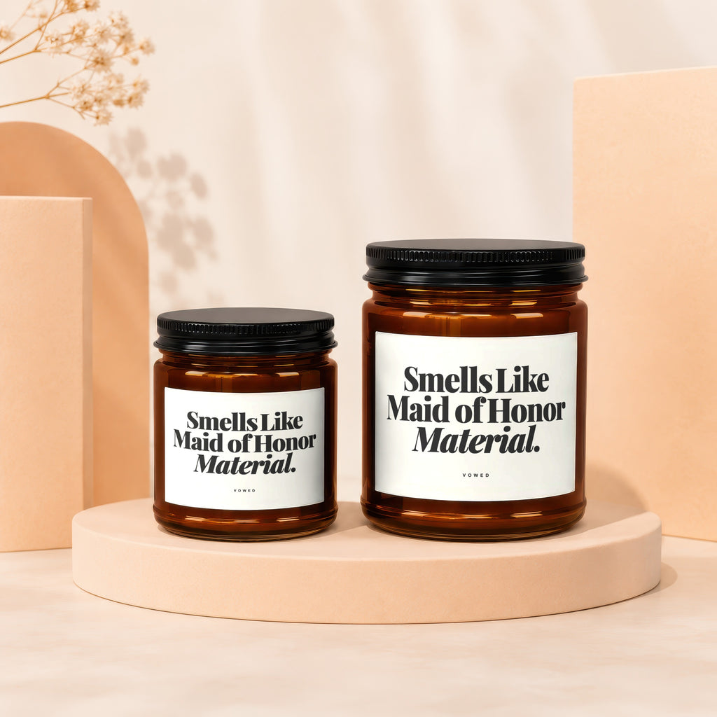 Two amber glass candles with black lids and white labels reading "Smells Like Maid of Honor Material."