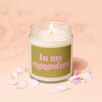 Lit candle with pink text "In my engaged era" on a green label, surrounded by petals.