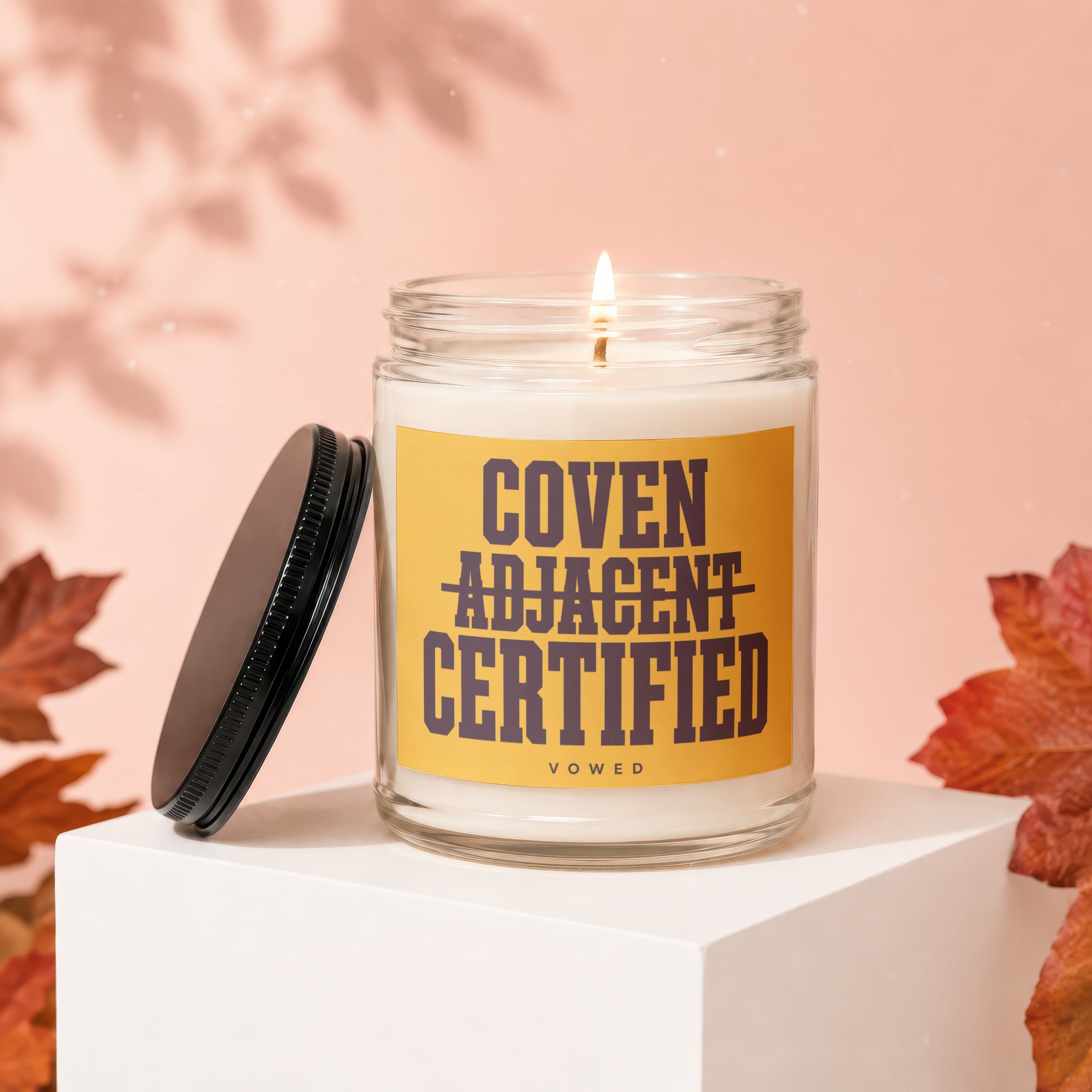 Lit candle in glass jar with "Coven Adjacent Certified Vowed" text on yellow label.
